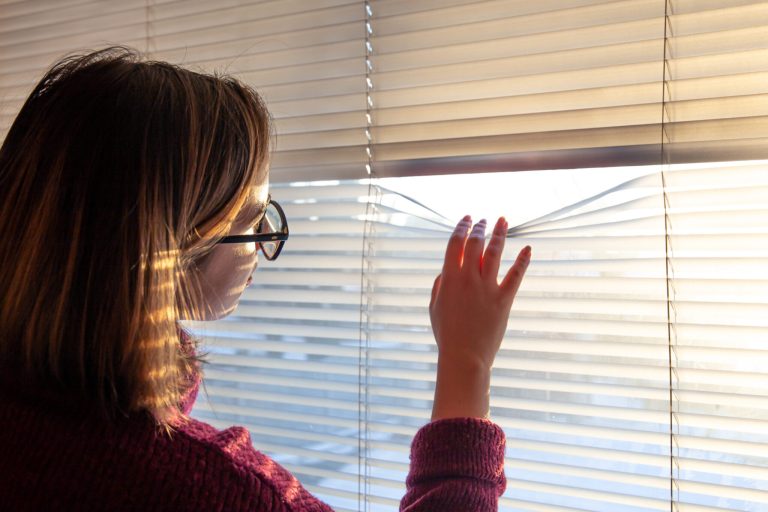 A woman looks through the blinds at the early morning sunlight.